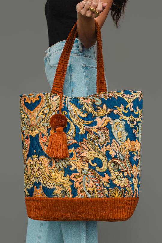 A person in blue jeans holds the Panache Apparel Collection’s Rust & Multicolored Patterned Tote, featuring an ornate blue, yellow, and orange paisley design, orange handles, and a tassel.