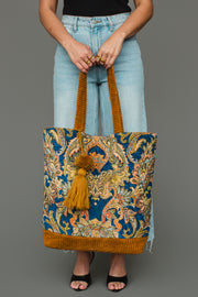 A person in light blue jeans and a black top holds the Mustard & Multicolored Patterned Tote by Panache Apparel Collection, featuring an ornate floral pattern, gold handles, and a gold tassel.
