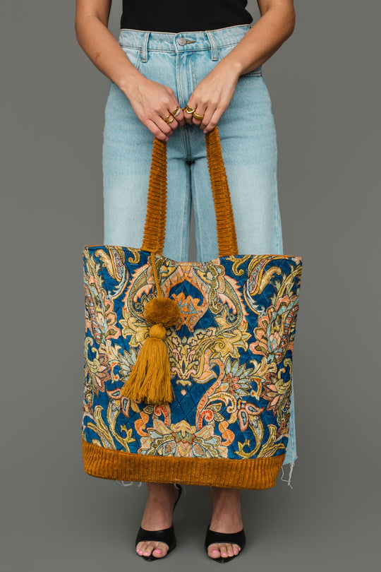 A person in light blue jeans and a black top holds the Mustard & Multicolored Patterned Tote by Panache Apparel Collection, featuring an ornate floral pattern, gold handles, and a gold tassel.