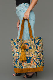 A person in jeans and black heels holds the Panache Apparel Collection’s Mustard & Multicolored Patterned Tote, featuring gold handles and a matching tassel, against a gray background.