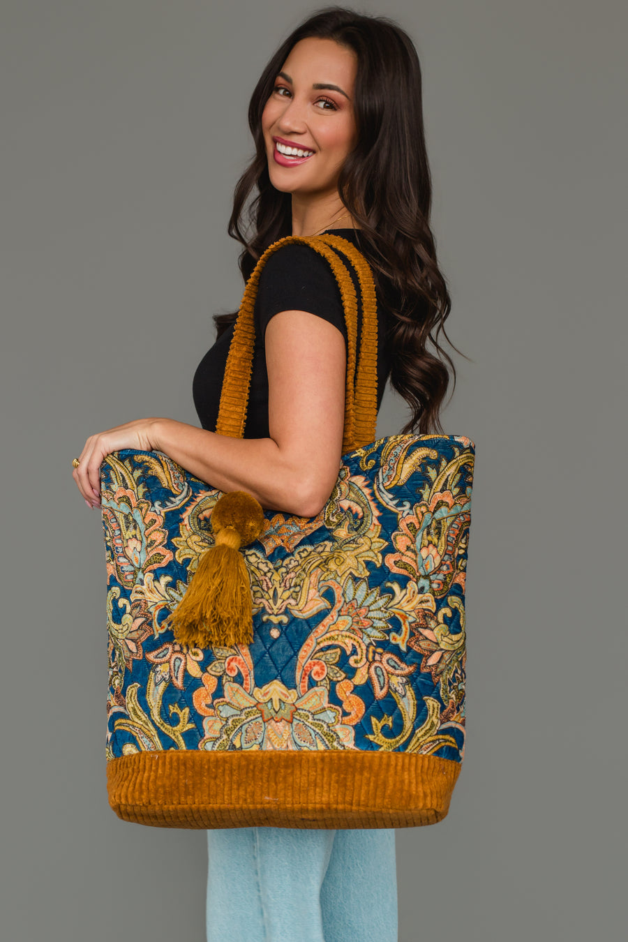 A woman with long dark hair, dressed in a black shirt and light jeans, smiles as she poses against a gray background, holding the Panache Apparel Collection Mustard & Multicolored Patterned Tote—featuring colorful designs, mustard yellow straps, and playful tassels.
