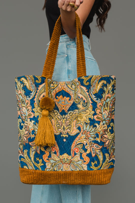 A woman wearing a black top and blue jeans holds the Mustard & Multicolored Patterned Tote from Panache Apparel Collection, featuring yellow and blue designs, brown textured handles, and a yellow tassel accent.