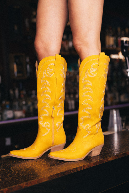 A person stands on a wooden bar counter in a dimly lit setting, wearing Panache Apparel Collection’s Yellow Embroidered Western Boots with decorative stitching. Bottles and glasses appear blurred in the background.