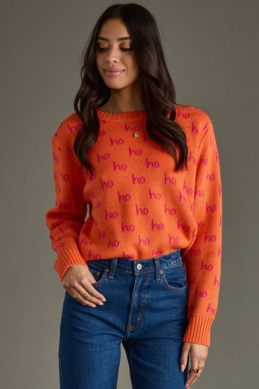 Woman wearing an orange sweater with 'ho ho ho' pattern and blue jeans against a gray background