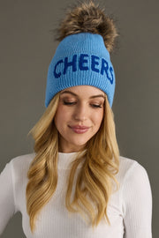 Woman wearing a blue beanie with 'CHEERS' text and fur pom-pom on a gray background
