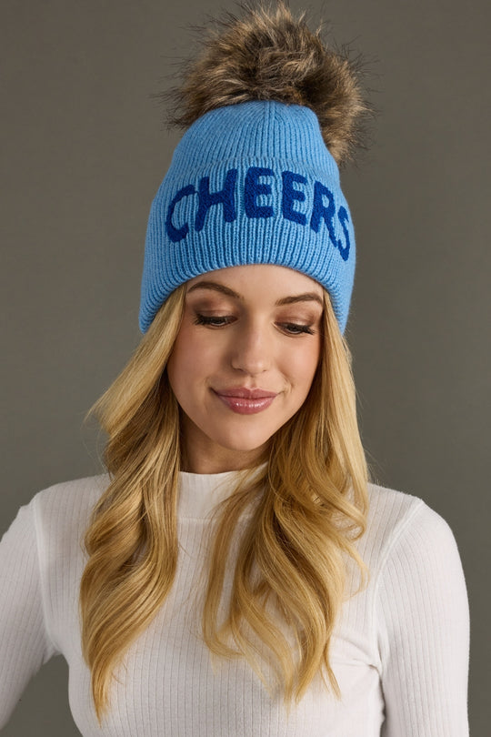 Woman wearing a blue beanie with 'CHEERS' text and fur pom-pom on a gray background