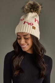 Woman wearing a cream knit beanie with red text and a fur pom-pom on a gray background