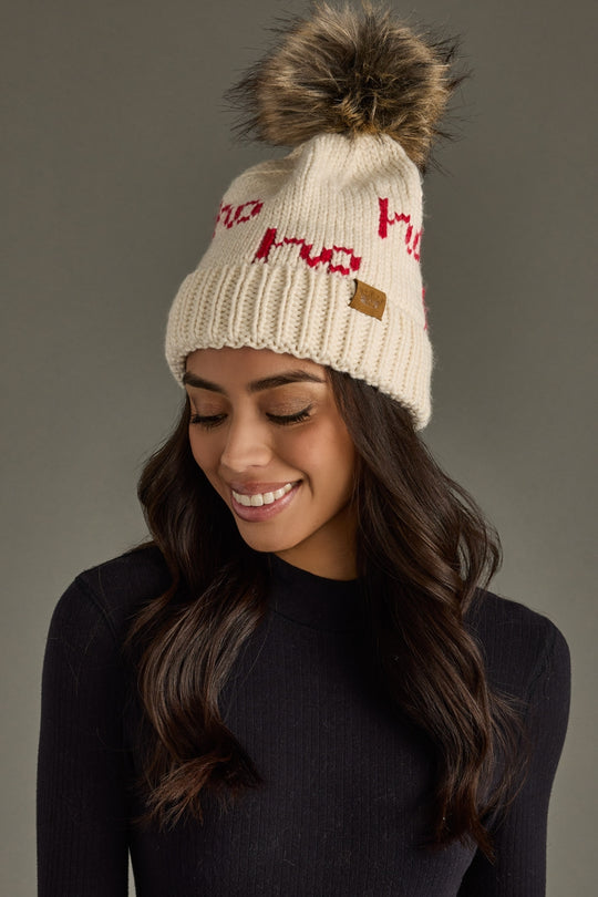 Woman wearing a cream knit beanie with red text and a fur pom-pom on a gray background
