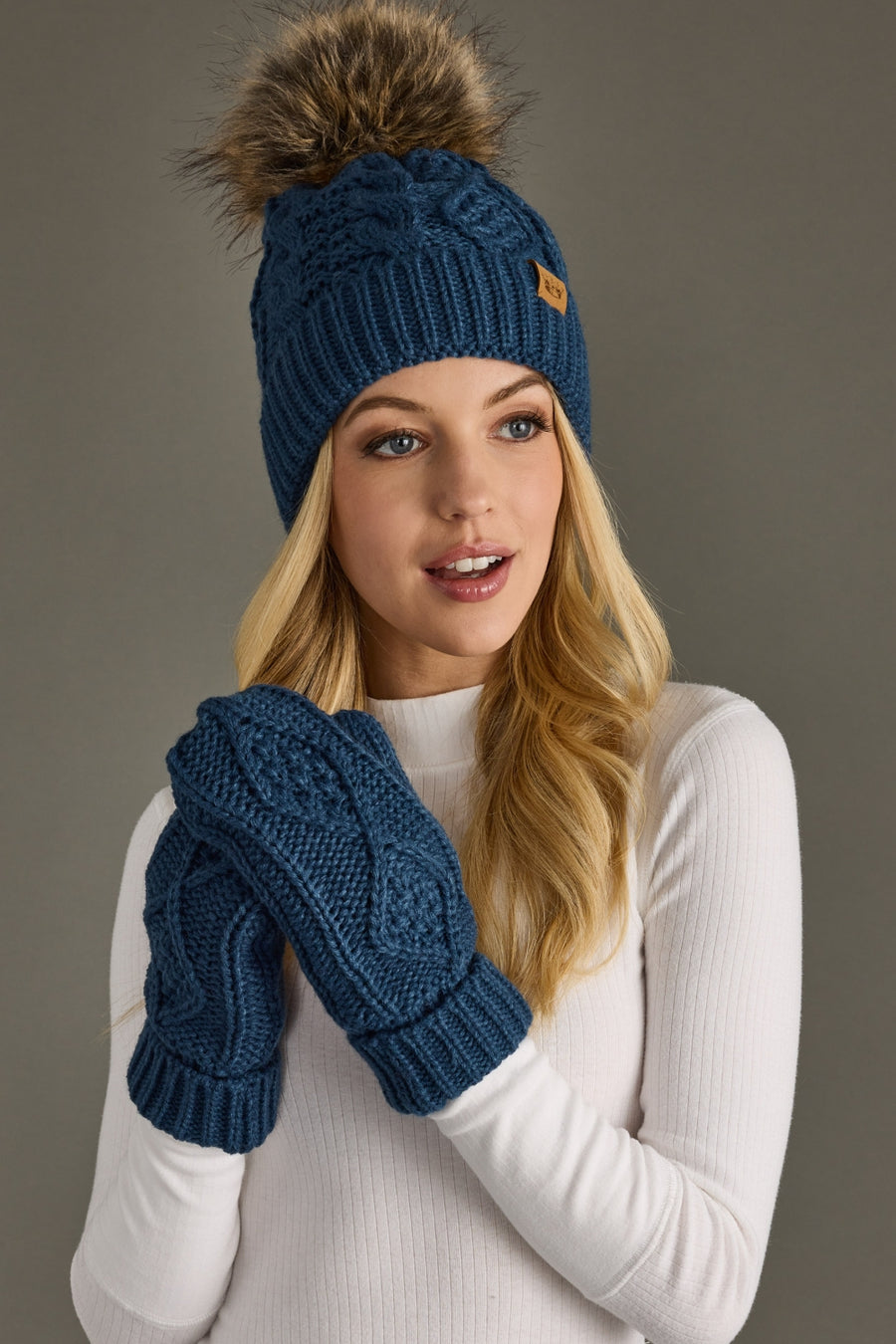 A woman poses in front of a grey background wearing Panache Apparel Collections Ramsey Mittens—cozy, fleece-lined, and cable knit in steel blue with a matching faux fur pom-pom hat and a white ribbed long-sleeve top.