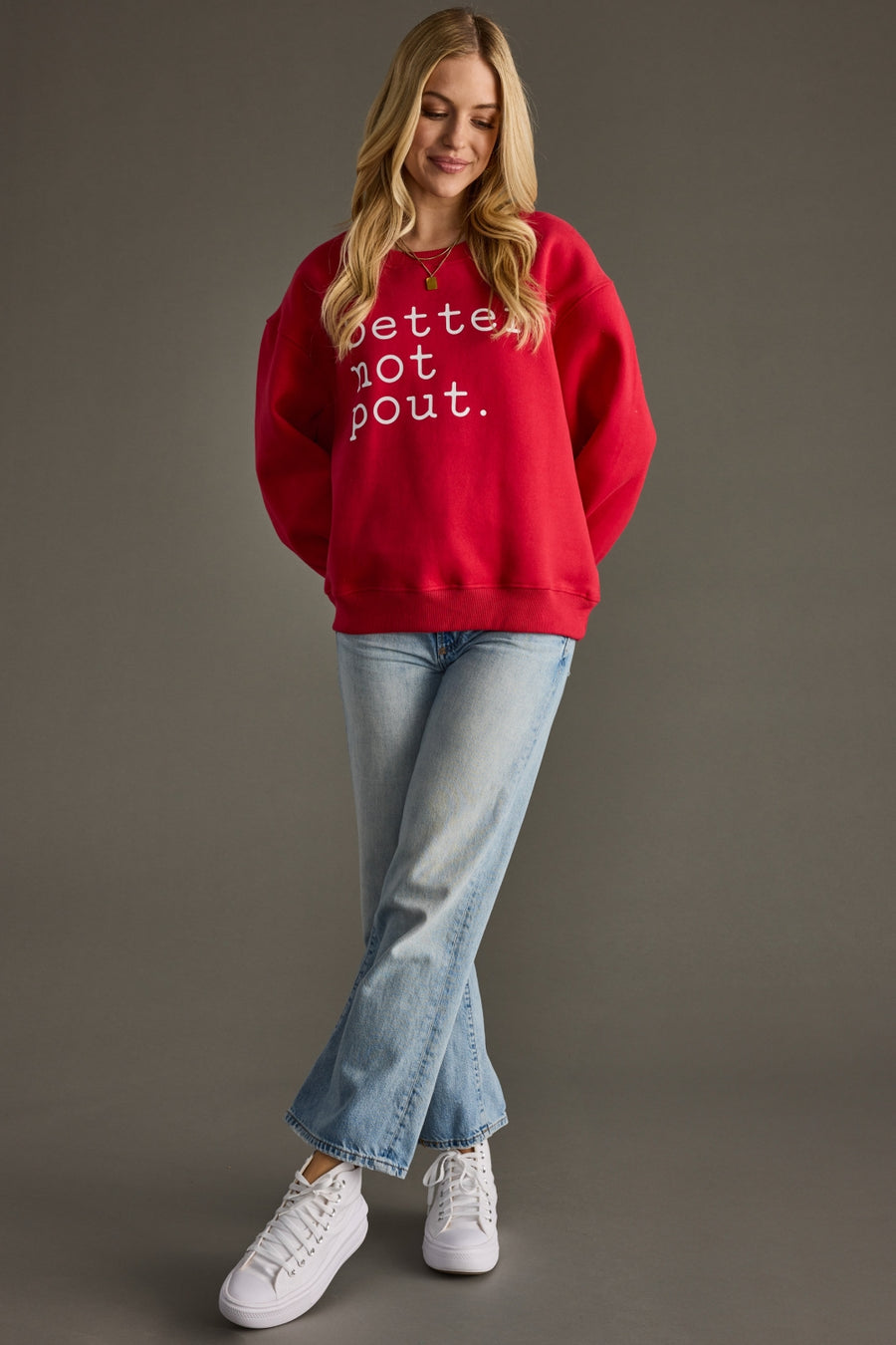 Woman wearing a red sweatshirt with text, blue jeans, and white sneakers on a gray background