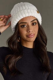 Woman wearing a white knit beanie with a hand adjusting it against a gray background