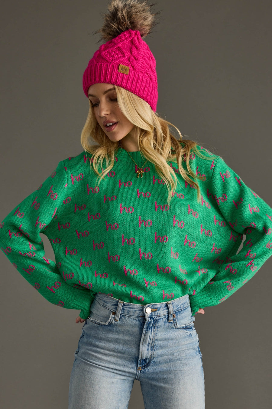 Woman wearing a green sweater with pink patterns, blue jeans, and a pink beanie against a gray background