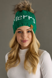 Woman wearing a green knit beanie with 'Merry' text and fur pom-pom against a gray background