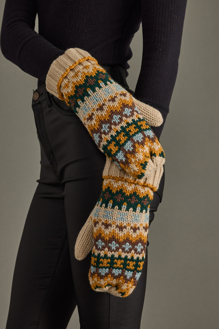 Pair of patterned mittens held by a person wearing black sleeves and pants against a gray background
