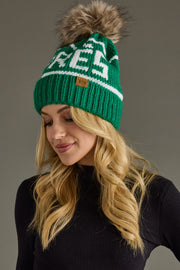 A woman with long blonde hair wears the Panache Apparel Collection "Hit The Slopes Pom Hat - Green & White," featuring a large faux fur pom and white stripe, paired with a black ribbed top against a plain gray background.