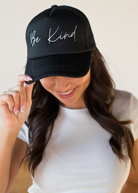 A woman with long brown hair wears a white short-sleeve top and smiles as she adjusts the Be Kind Trucker Hat by panacheaccessories—featuring "Be Kind" in white script and an adjustable snapback for comfort.