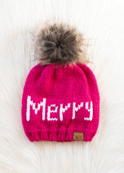 Pink knit beanie with 'Merry' text and fur pom-pom on a light background