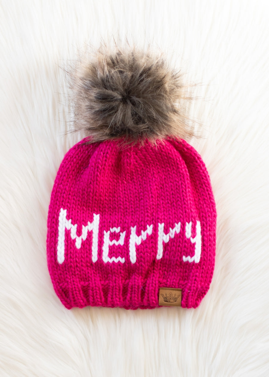 Pink knit beanie with 'Merry' text and fur pom-pom on a light background