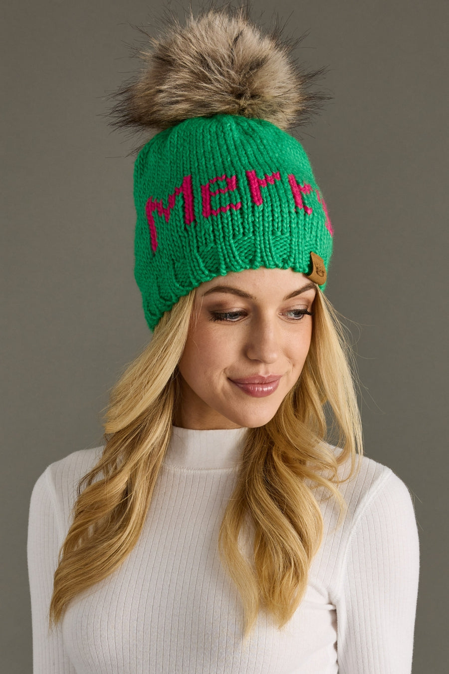 A woman with long blonde hair wears the Panache Apparel Collection Merry Pom Hat in bright green, featuring a large faux fur pom-pom and red "Merry" lettering, paired with a white ribbed turtleneck as she smiles softly.