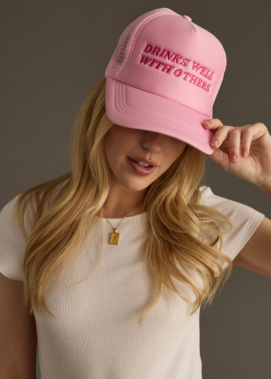 A woman in a cream ribbed top wears a bright pink trucker hat with bold text that reads “DRINKS WELL WITH OTHERS.” She’s tilting the brim down slightly, giving the whole shot a playful, cheeky vibe.