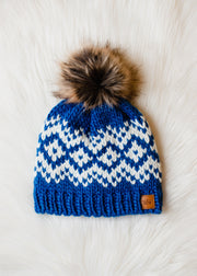 Blue knitted beanie with white pattern and brown faux fur pom-pom on a white background