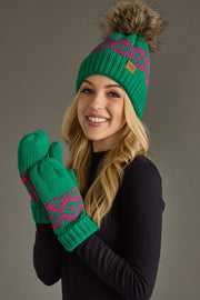A smiling woman with long blonde hair wears the After Hours Pom Hat by Panache Apparel Collection, featuring a green knit design with a brown faux fur pom-pom, paired with matching green fleece-lined mittens and a black sweater.