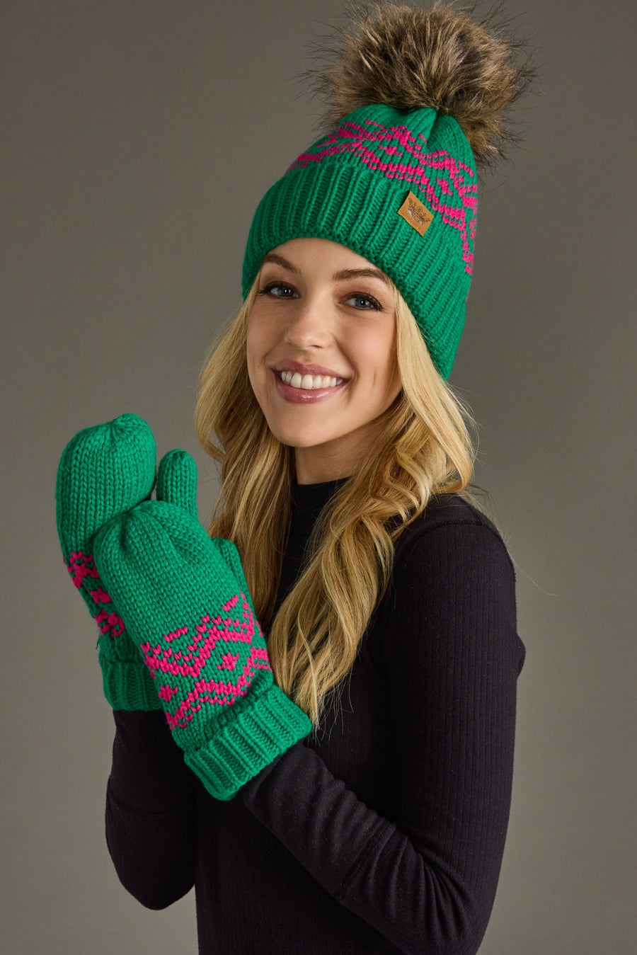 A smiling woman with long blonde hair wears the After Hours Pom Hat by Panache Apparel Collection, featuring a green knit design with a brown faux fur pom-pom, paired with matching green fleece-lined mittens and a black sweater.