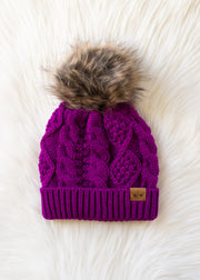 Purple knit beanie with fur pom-pom on a white fluffy surface