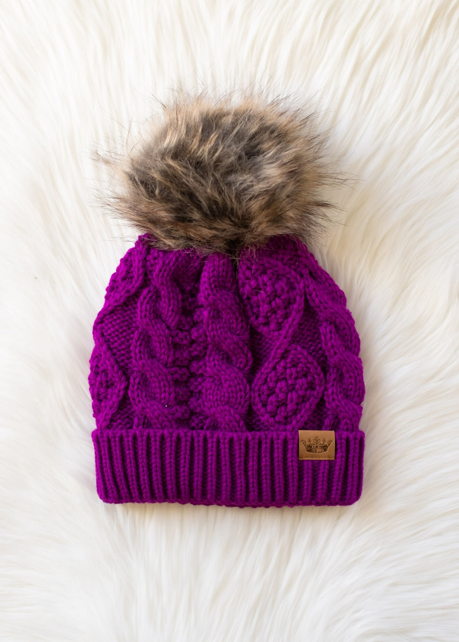 Purple knit beanie with fur pom-pom on a white fluffy surface