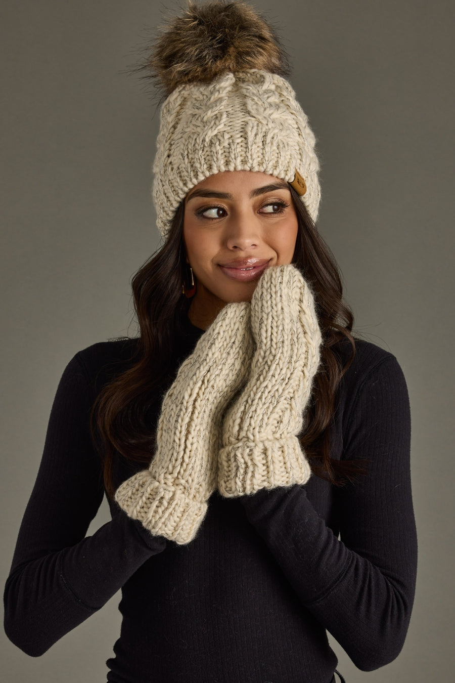 Woman wearing a cream knit beanie with a pom-pom and matching gloves against a gray background