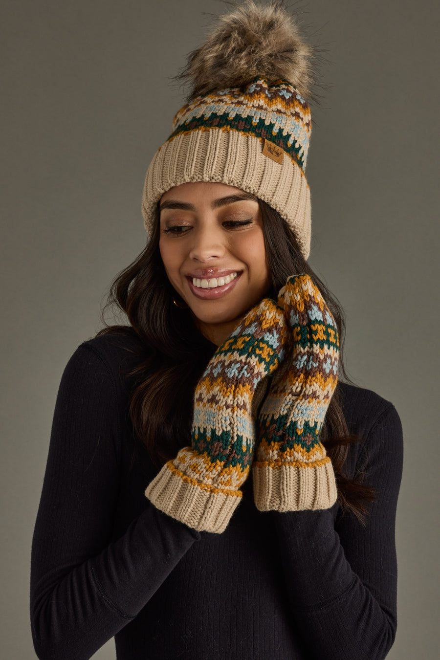 Woman wearing a knitted hat and mittens with a fur pom-pom against a gray background