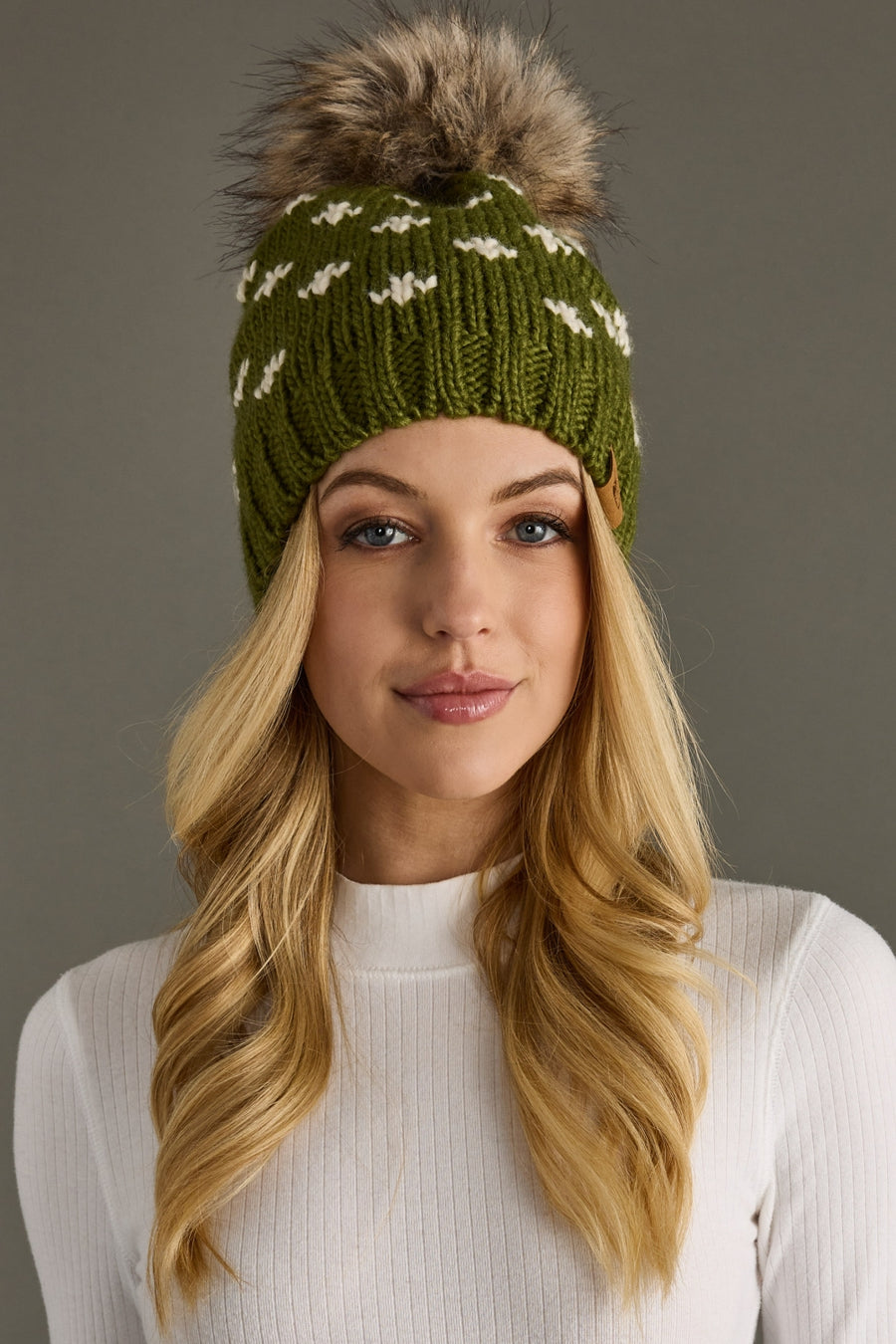Woman wearing a green knit beanie with a fur pom-pom against a gray background