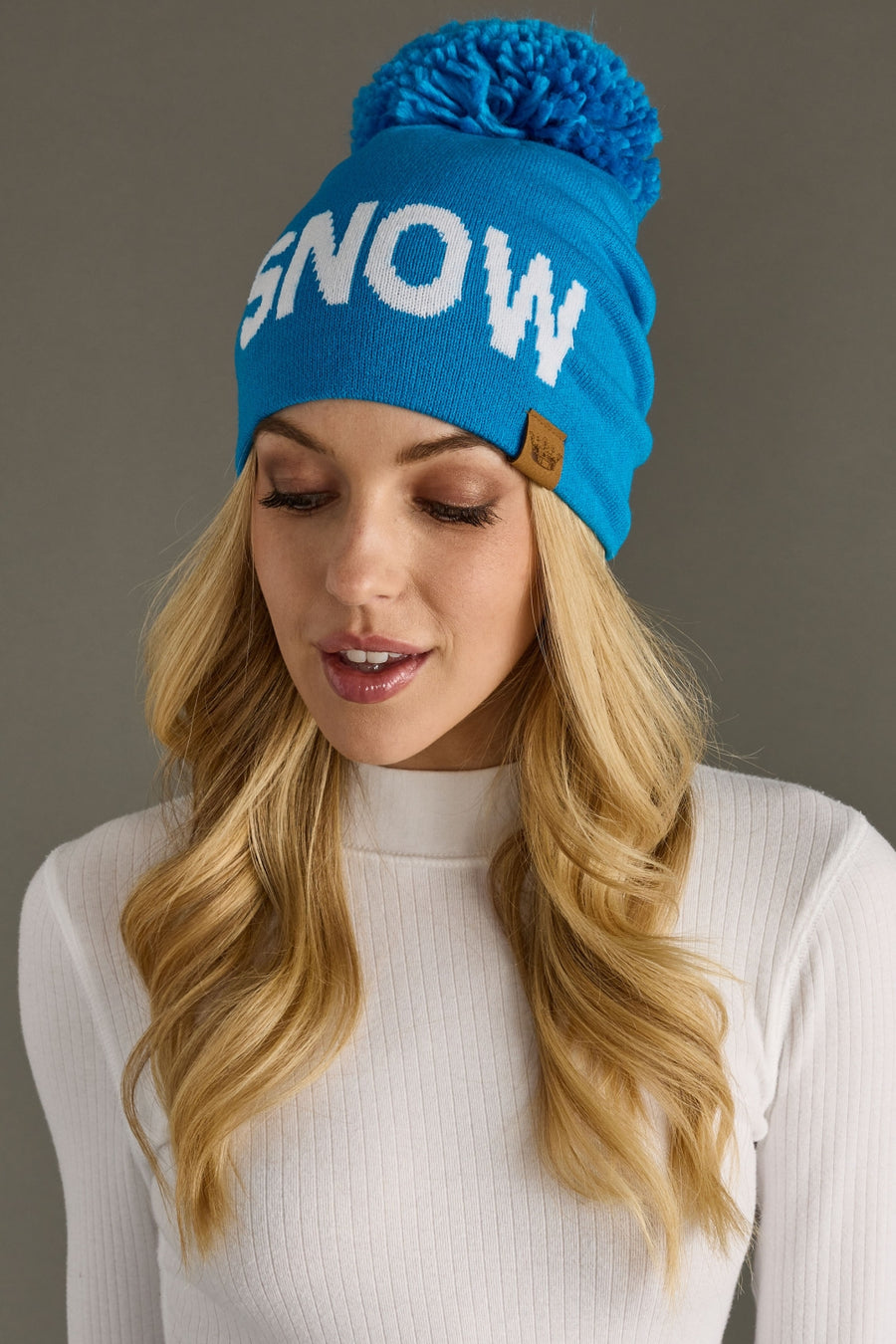 Woman wearing a blue beanie with 'SNOW' text and pom-pom on a gray background