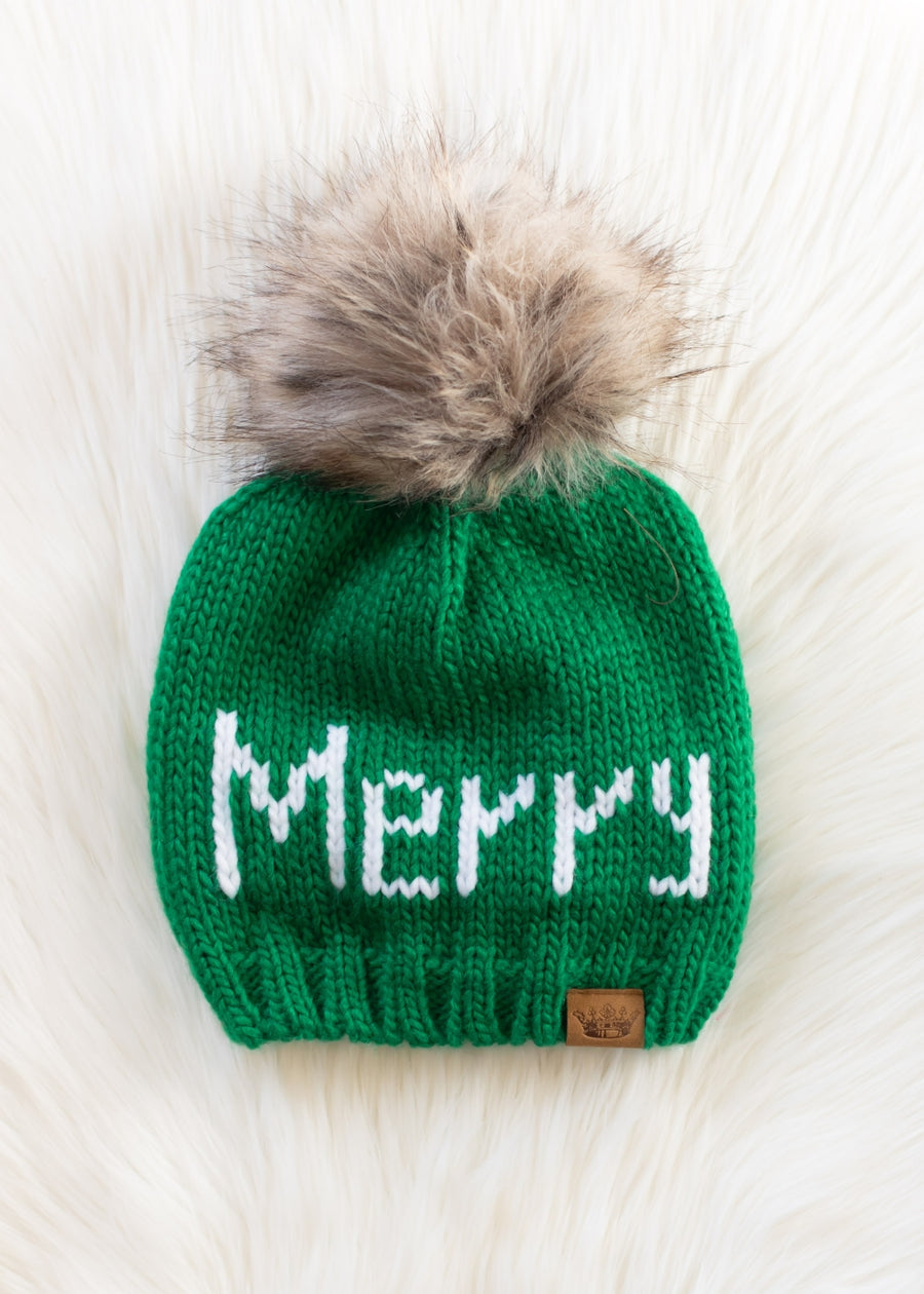 Green knit beanie with 'Merry' text and fur pom-pom on a light wooden surface