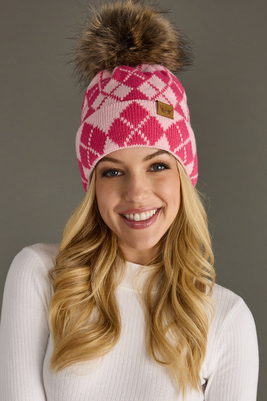 Woman wearing a pink and blush patterned beanie with a fur pom-pom on a gray background