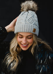 A woman with long blonde hair smiles while looking slightly to the side, wearing a light gray knit beanie with a faux-fur pom-pom and a small leather patch on the front. She’s dressed in a dark, glossy winter jacket, posed against a plain dark background that highlights her warm expression and cozy cold-weather style.