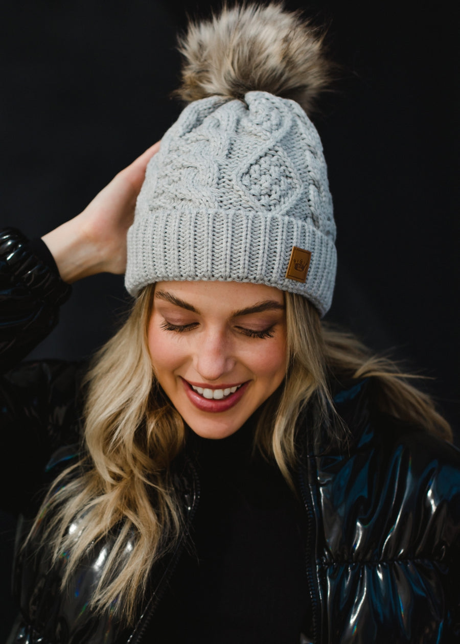 A woman with long blonde hair smiles while looking slightly to the side, wearing a light gray knit beanie with a faux-fur pom-pom and a small leather patch on the front. She’s dressed in a dark, glossy winter jacket, posed against a plain dark background that highlights her warm expression and cozy cold-weather style.