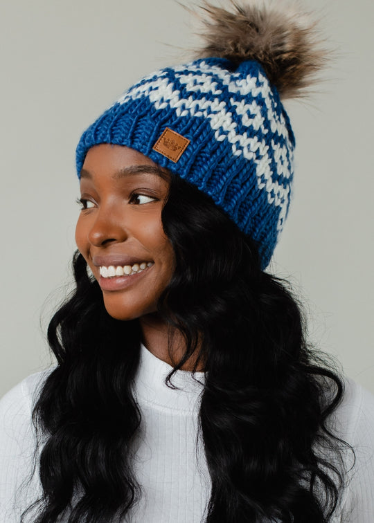 Woman wearing a blue and white patterned beanie with a brown pom-pom on a neutral background