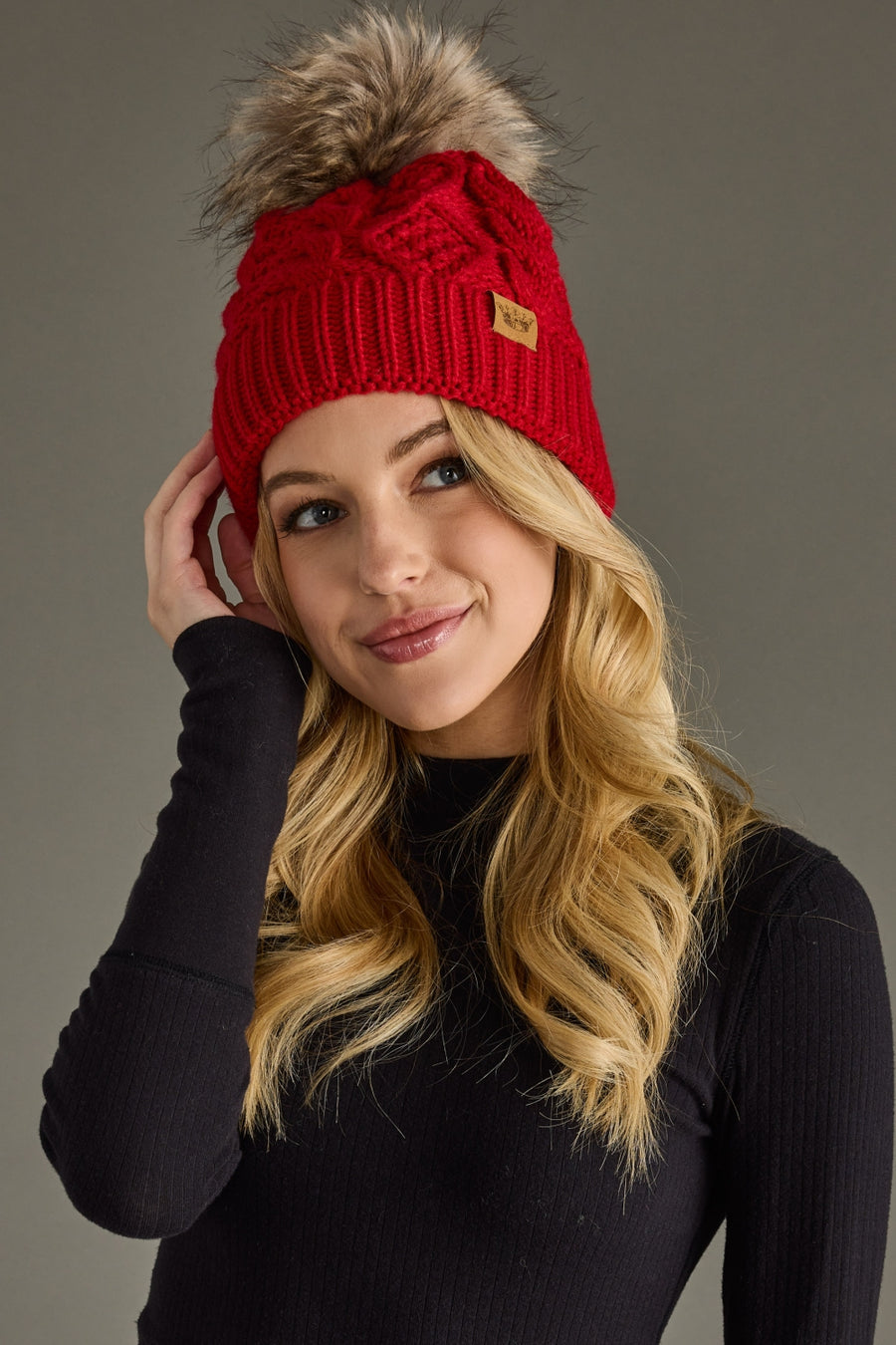 A woman with long blonde hair, wearing the Panache Apparel Collection Red Cable Knit Pom Hat, which features a faux fur pom, and a black long-sleeve top, smiles softly while touching her hat against a plain gray background.
