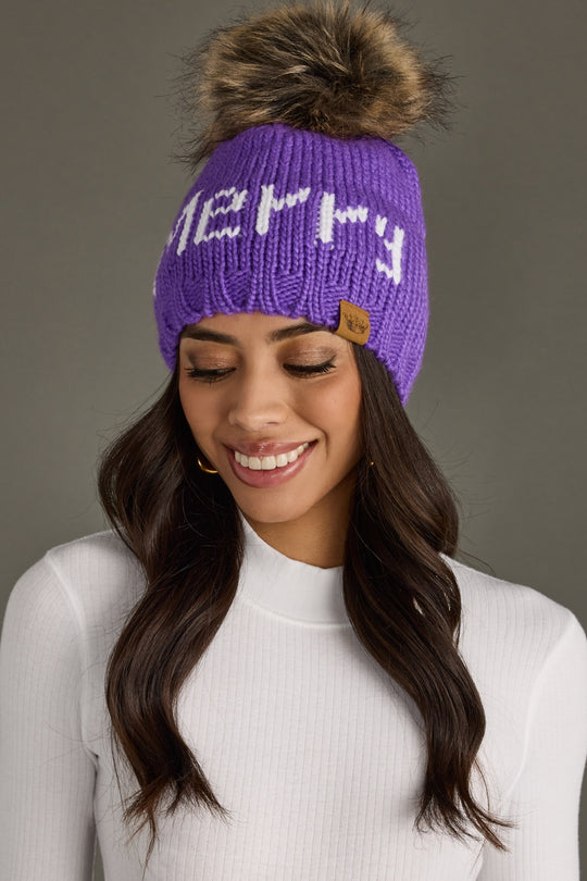 Woman wearing a purple knit beanie with a fluffy pom-pom on a gray background