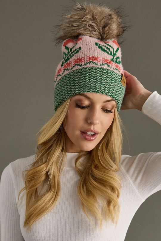 Woman wearing a green and pink knitted hat with a fur pom-pom against a gray background