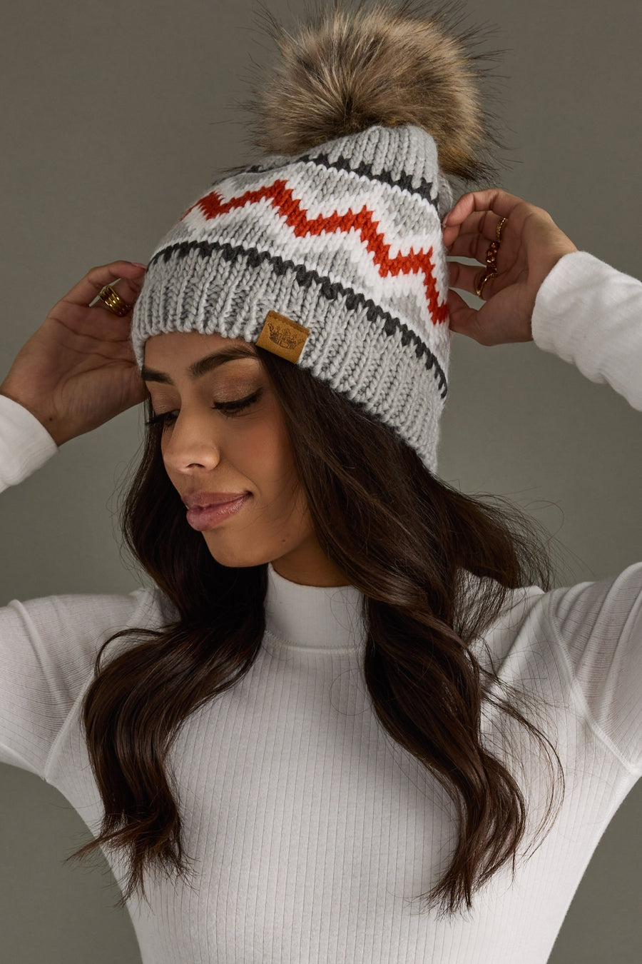 Woman wearing a gray knit beanie with a pom-pom and red zigzag pattern against a neutral background