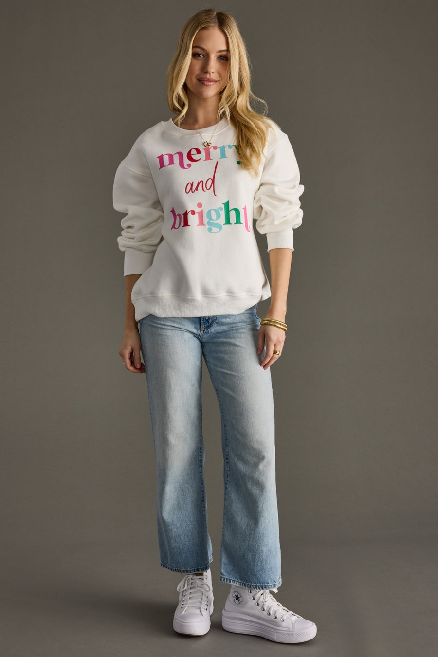 Woman wearing a white sweatshirt with colorful text and blue jeans on a gray background