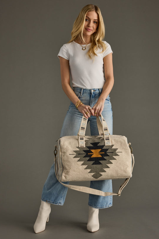 Woman holding a beige duffel bag with geometric patterns against a gray background