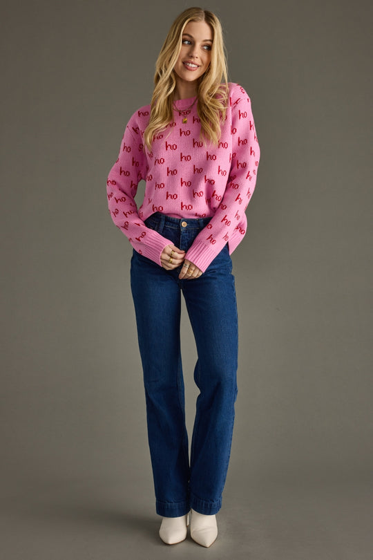 Woman wearing a pink sweater with red text and blue jeans on a gray background