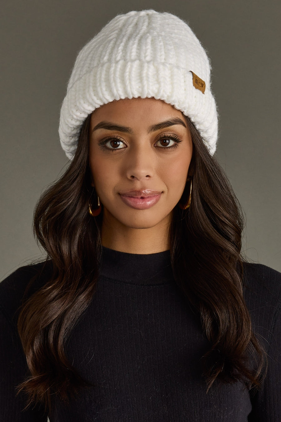 Woman wearing a white knit beanie with a brand logo on a gray background