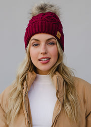 Woman wearing a red knit beanie with a fur pom-pom against a plain background