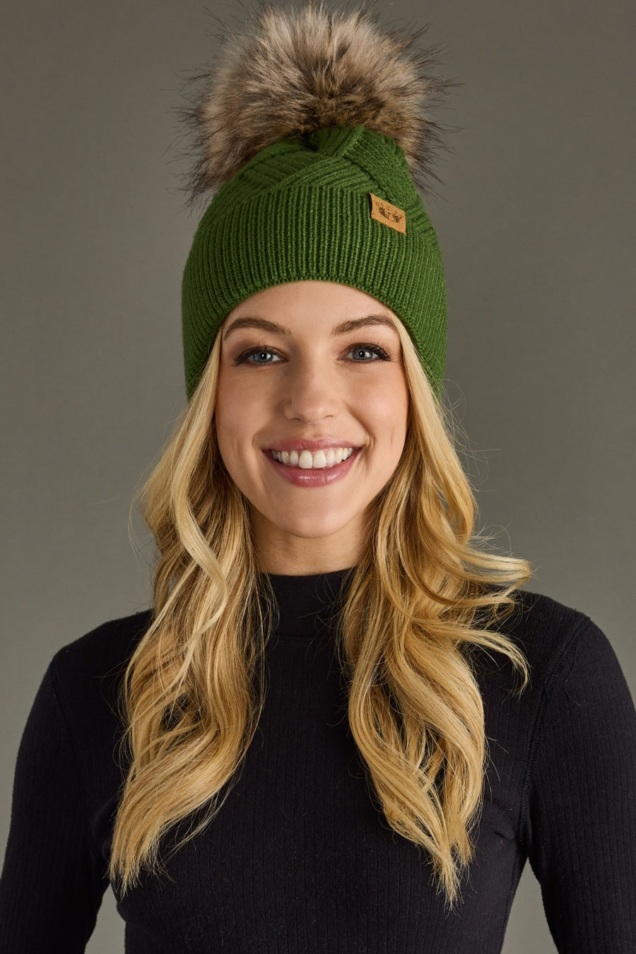 Woman wearing a green knit beanie with a fur pom-pom against a gray background