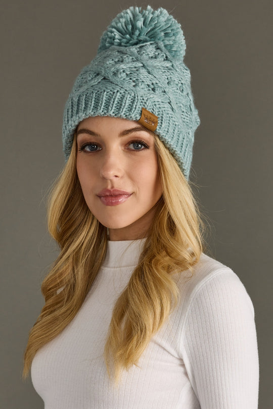 Woman wearing a light blue knit beanie with a pom-pom against a gray background