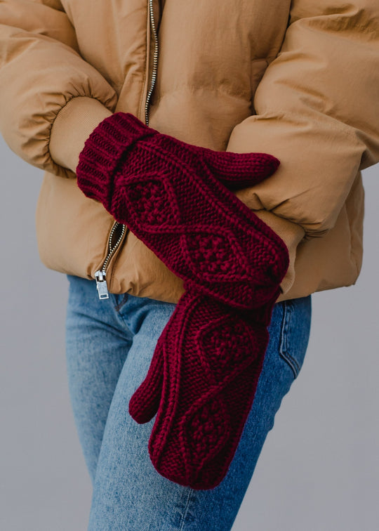 Person wearing red knitted mittens with a beige coat and blue jeans on a gray background